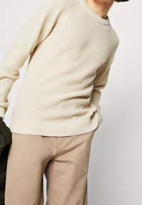 Beige strukturerad stickad tröja med rundad halsringning och ribbad nederkant, i kombination med ljusbeige byxor. Inga påtagliga mönster eller detaljer synliga.