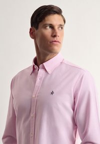 Camisa de botón de color rosa claro, hecha de algodón, con cuello extendido y un pequeño logo bordado en el lado izquierdo del pecho. Textura de tela suave.