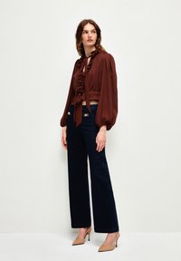 Blusa marrón cropped con detalles fruncidos y cierre con lazo, combinada con vaqueros de pernera ancha en color marino y zapatos de tacón beige. Fondo liso.