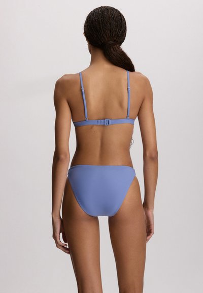 Γυναίκα με πλεξούδες, τα μαλλιά της δεμένα πίσω, φοράει ανοιχτό μπλε μπικίνι, εμφανίζεται από την πίσω πλευρά με φόντο ένα απλό υπόβαθρο.