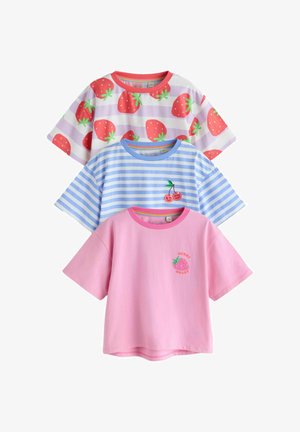 Mosolygó fiatal lány hosszú barna hajjal, fehér és levendulalila csíkos ingben, amelyet nagy piros eprek díszítenek, bézs háttér előtt.