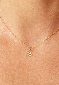 Halskette in Goldtönen mit dünner Kette und Anhänger in Form des Kleinbuchstabens 'w'. Glatte Oberfläche mit minimalistischem Design.