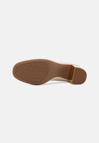Semelle d'une chaussure à talon beige avec un motif de semelle texturé répétant le logo et un talon bloc sur fond blanc uni.