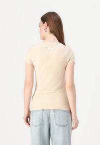 T-shirt bege de manga curta com um design ajustado. Apresenta um pequeno logo branco na parte superior das costas. Textura suave e lisa. Combinado com jeans azuis claros.