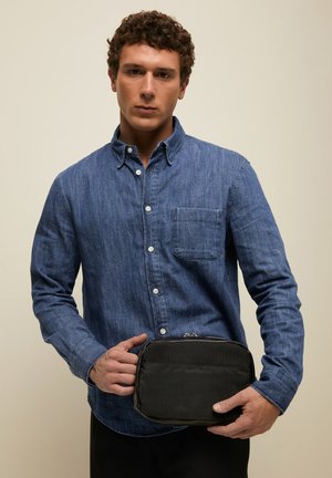 Mees, kes kannab sinist nööbitavat särki, hoiab väikest musta lukuga kotikest ühetaolise beeži tausta ees.