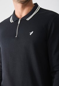 Sort langærmet polo-shirt med lynlås-krave, prydet med hvide striber og et lille hvidt fuglelogo på brystet. Blød stofstruktur.