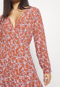 Blommig klänning i rostorange och ljusrosa med V-ringning, långa ärmar och omslagsdesign. Har ett mönstrat tyg med en texturerad yta.
