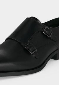 Chaussure habillée en cuir noir avec double bride à boucle carrée en métal, bout pointu et talon bas sur fond blanc.