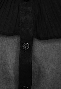 Camicetta nera con bottone a pressione che presenta uno yoke a pieghe testurizzato, tessuto morbido e bottoni neri. Il design è semplice con un taglio classico.