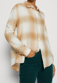 Camisa de botones de manga larga a cuadros en tonos beige y blanco con cuello. Se muestra combinada con pantalones de pana en color teal, destacando un ajuste relajado y una textura suave.