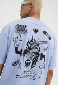 Person som har på seg en lys blå skjorte med svart grafikk av en animert takeout-boks og spisepinner, med ordene «STAY CHILL» og «BETTER TOMORROW.»