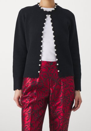 Cardigan noir avec bordure en perles sur un top blanc côtelé, associé à un pantalon en satin rouge avec un motif floral damassé sombre.