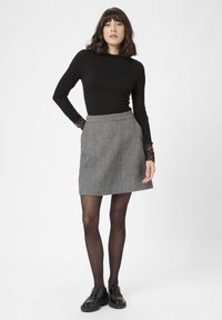 Vrouw die staat met hand in zak, gekleed in een zwart longsleeve shirt met kanten manchetten, een grijze patroonrok, zwarte panty en zwarte schoenen op een witte achtergrond.