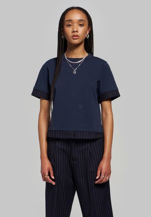 Jeune femme aux longues tresses, portant un haut bleu marine à plusieurs couches et un pantalon bleu marine à fines rayures, accessoirisée de bijoux argentés, debout devant un fond gris.
