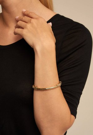 Bracelet en or avec un design torsadé et des accents texturés. Porté au poignet sur un fond de chemise noire.