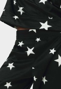 Zwart fleeceloungewear set met witte sterrenpatronen; de top heeft een losse pasvorm en korte mouwen, terwijl de broek een elastische tailleband heeft.