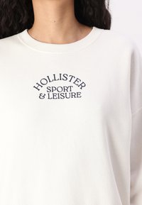 Bijela dukserica s okruglim izrezom, s vezom "HOLLISTER SPORT & LEISURE" u tamnoplavoj boji. Mekani pamučni materijal s blagom teksturom.