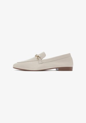 Mocassins en cuir beige avec un talon plat, arborant une boucle dorée décorative sur le dessus et un design minimaliste. Texture lisse et silhouette épurée.