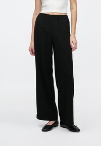 Pantalon noir à jambes larges en tissu doux avec une ceinture élastique. Le design présente une coupe ample et un style minimaliste, associé à des ballerines noires.