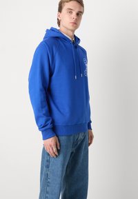 Sudadera azul de tejido suave, con capucha ajustable, puños acanalados y un diseño gráfico en el pecho. Combinada con jeans de mezclilla claros.