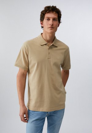 Uomo con capelli ricci che indossa una polo beige a maniche corte e jeans azzurri, in piedi davanti a uno sfondo chiaro e uniforme.