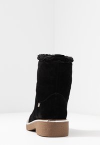 Svart mocka ankelstövel med brun gummisula och lammullskant, visad från bakvinkeln på vit bakgrund.