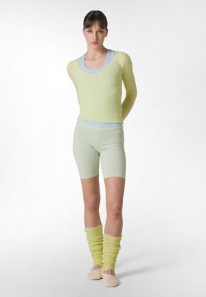 Top lung cu mâneci lungi, texturat, de culoare galben deschis, peste un maieu de culoare albastră deschisă, asortat cu pantaloni scurți verzi pastel și glezne asortate, pantofi de balet roz deschis.