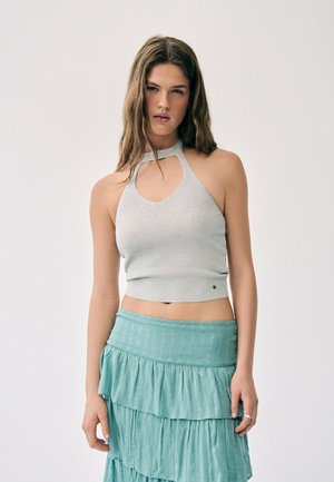 Jeune femme portant un haut dos nu gris clair et une jupe bleue claire à volants, debout devant un fond blanc uni.