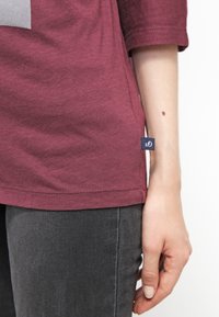 Burgundy cotton T-shirt with three-quarter sleeves, side slit, and small blue fabric label at the hem. Paired with dark gray pants.