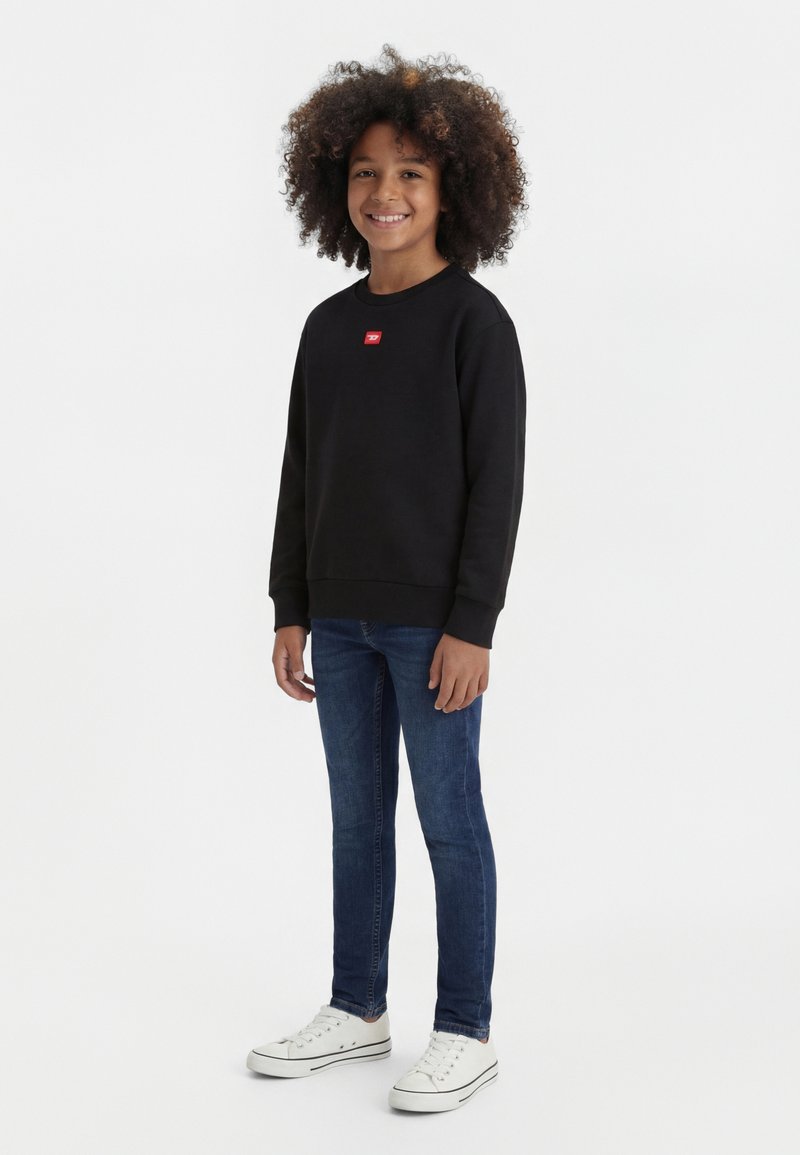 Usměvavé dítě s kudrnatými vlasy má na sobě černou mikinu, modré džíny a bílé tenisky, stojící před jednobarevným bílým pozadím.