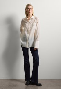 Camicia bianca a maniche lunghe con chiusura a bottoni, taschino sul lato sinistro del petto e una texture liscia, abbinata a jeans flared scuri e scarpe nere.