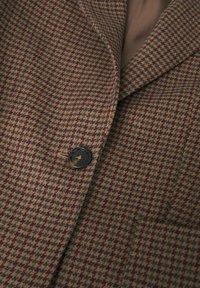 Gros plan sur un blazer à motif pied-de-poule marron, vert et violet, avec un bouton noir et un revers visible.