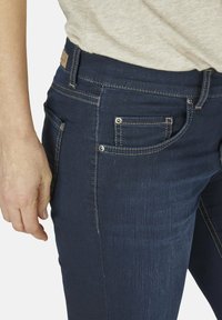 Tmavě modré džíny z denimu s viditelným šitím, předními a zadními kapsami a kovovými akcenty. Tkanina má hladký, mírně texturovaný povrch.