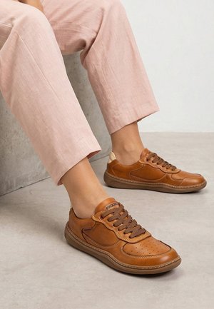 Brune lædersneakers med mørkebrune snørebånd, båret med lyserøde croppede bukser mod en neutral gulv- og vægbaggrund.