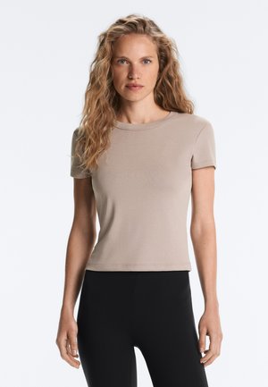 Femme aux longs cheveux blonds bouclés portant un t-shirt beige ajusté à manches courtes et un pantalon noir, debout devant un fond blanc uni.