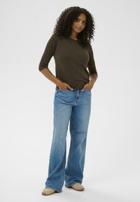 Brązowa, dopasowana bluzka z długim rękawem zestawiona z jasnoniebieskimi spodniami dzwonami. Modelka stoi z rękami w kieszeniach, ubrana w beżowe buty. Jednolity tle.