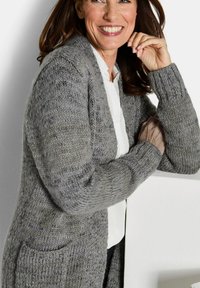 Lächelnde Frau mit braunem Haar, die eine graue Strickjacke über einem weißen Shirt trägt und mit verschränkten Armen auf einer weißen Fläche lehnt.