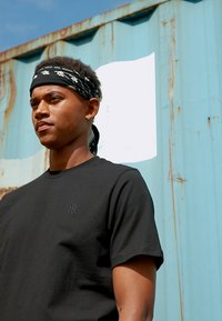Black cotton t-shirt with short sleeves, featuring a small logo on the chest. The model wears a black patterned bandana against a blue background.