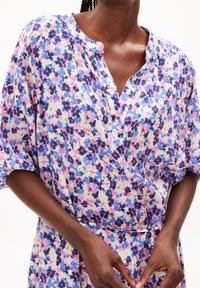 Hame floral dengan cetakan, menggunakan kain ringan, menampilkan leher V, lengan pendek, dan pita di pinggang. Warna mencakup merah muda, ungu, dan kuning di latar belakang yang cerah.