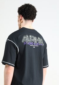 T-shirt noir avec un imprimé graphique dans le dos portant l'inscription "ALPHA" en finition brillante. Finitions en fil argenté sur les manches.