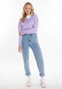 Levandulový pletený cardigan s výstřihem do V a knoflíkovým upevněním, v kombinaci s vysokými světle modrými džínami a pastelovými sneakers, na jednobarevném pozadí.