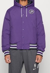 Lila puffjacka med huva, med vita dragsko, tryckknappar och randiga mudd. Inkluderar en basketlogo på bröstet.