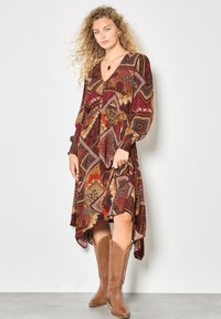 Langärmliges Kleid mit tiefem V-Ausschnitt, das ein mehrfarbiges geometrisches Muster in erdigen Tönen aufweist. Kombiniert mit hellbraunen Cowboy-Stiefeln.