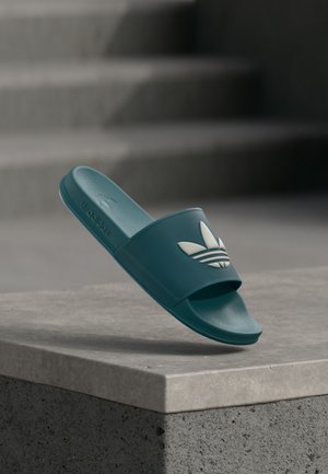 Sandale slide Adidas bleu sarcelle avec logo trèfle blanc, montrée en l’air au-dessus d’un rebord en béton et d’un fond d’escalier flou.