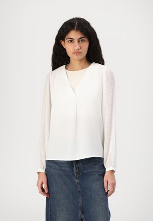 White blouse with a V-neck, sheer dotted sleeves, and a smooth bodice. Paired with a denim skirt, showcasing a casual look.