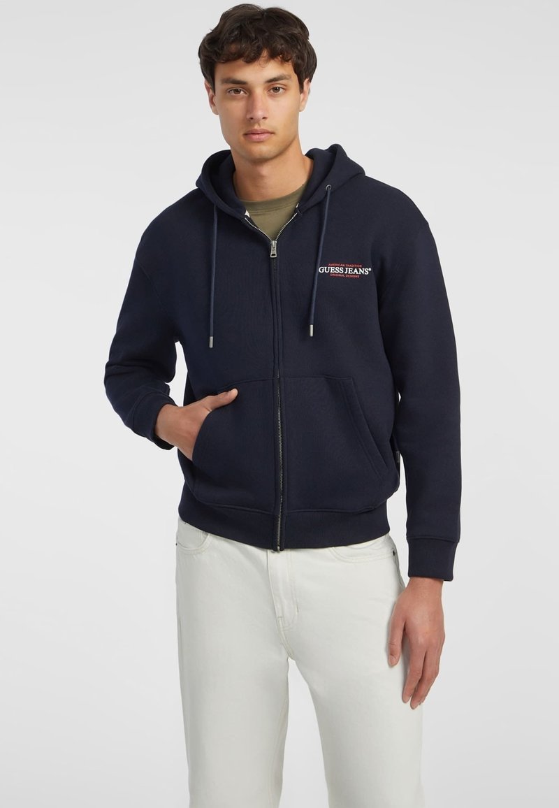 Marineblaue Fleecekapuzenjacke mit Reißverschluss, Fronttaschen, Kordelzügen und Logodetails auf der Brust. Kombiniert mit weißen Jeans.