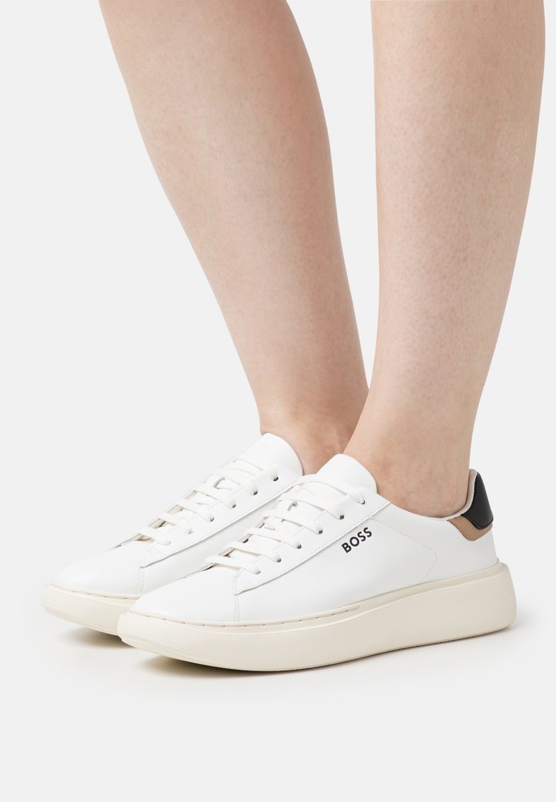 Baskets basses blanches avec le logo « BOSS » sur le côté, semelles beige, portées pieds nus sur un fond clair uni.