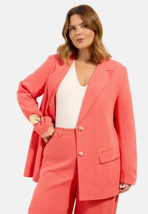 Frau trägt korallenroten Blazer und passende weite Hose mit weißem V-Ausschnitt-Top, posiert mit einer Hand in der Hüfte vor weißem Hintergrund.