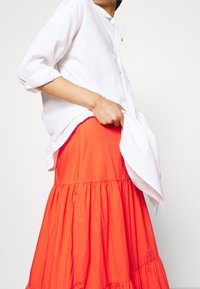 Henkilö, joka on pukeutunut löysään valkoiseen nappipaitaan ja kirkkaan oranssiin kerrokselliseen hameeseen, pitää kevyesti paitaa vyötäröllä yksinkertaista taustaa vasten.