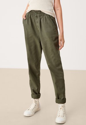 Osoba nosící olivově zelené kalhoty s vysokým pasem a knoflíkovým zapínáním, ohrnuté lemy, boční kapsy, v kombinaci s bílými vysokými teniskami a béžovým tílkem.